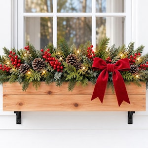 May include: A rectangular light-toned wooden window box filled with evergreen branches, pinecones, and clusters of bright red berries. A large red velvet bow is attached to the right side, and small warm white lights are woven throughout.