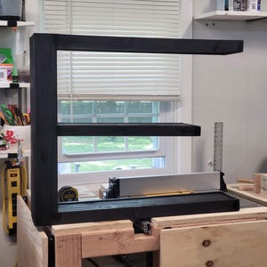 May include: A black wooden shelf unit with three levels. The unit is positioned above a wooden workbench with a silver saw. The background includes a window with white blinds and other workshop items.