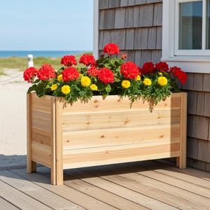 May include: A rectangular wooden planter box filled with vibrant red and yellow flowers. The planter is made of light-colored wood and sits on a wooden deck. The background shows a beach and the ocean.