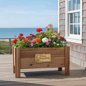 May include: A wooden rectangular planter box filled with colorful flowers. The box has a decorative nameplate that reads "The Smith". The planter is on a wooden deck with a view of the ocean.