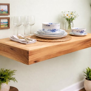 May include: A floating wooden shelf with a natural finish, displaying a set of white and blue floral patterned dishes, wine glasses, and silverware. The shelf is mounted on a light-colored wall, with small framed landscape paintings above it. A vase of daisies and a potted plant are also visible.