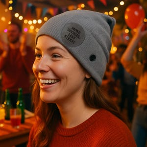 May include: A gray beanie with a circular patch that reads "MORE COOKIES, LESS STRESS." The beanie has a black button detail and is worn by a person smiling. The background is blurred with party lights and people.