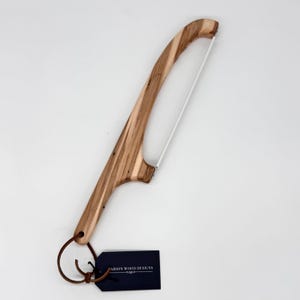 May include: A wooden cheese slicer with a curved handle and a thin, white wire blade. The handle is made of light brown wood with dark brown grain patterns. A dark blue tag with white text is attached to the handle.