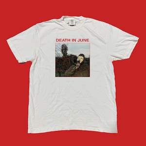 Può includere: T-shirt bianca con la scritta rossa "DEATH IN JUNE" sopra un'immagine incorniciata. L'immagine presenta una persona con una maschera che tiene in mano una tromba, sullo sfondo di rami e fogliame contorti.