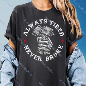 May include: Dark gray t-shirt with the text "ALWAYS TIRED NEVER BROKE" in a circular design. The graphic features a skeletal hand holding money, with red lightning bolts on either side. A casual, comfortable tee.