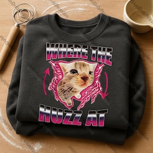 Op de afbeelding: Donkergrijze sweatshirt met een katje-afbeelding. De afbeelding bevat de tekst "WHERE THE HUZZ AT" in een roze en witte gradiënt, met een roze bliksemontwerp rond het katje. Het katje is lichtbruin en wit.