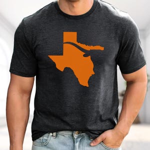 May include: A charcoal grey short-sleeve t-shirt featuring a solid orange graphic of the state of Texas on the chest. The state outline includes a stylized, curved horizontal band across the upper portion, rendered in the same orange color.