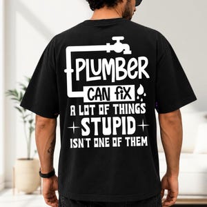 May include: A black short-sleeved t-shirt featuring white graphic text on the back that reads: PLUMBER CAN fix A LOT OF THINGS STUPID ISN'T ONE OF THEM, accompanied by a white faucet icon and a water drop symbol above the text.
