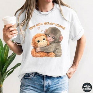 May include: White t-shirt with a graphic of two monkeys embracing, accompanied by the text "you are never alone" in orange. The monkeys are brown and orange. The shirt is worn by a person wearing blue jeans.