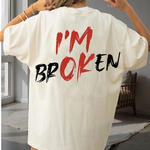 May include: Cream-colored t-shirt with the words "I'M BROKEN" printed on the back in red and black. The text has a distressed, brushstroke style. The shirt is a casual, oversized fit.