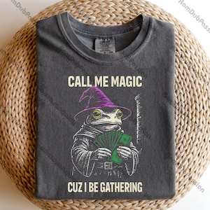 May include: Dark gray t-shirt with a graphic of a frog wearing a purple wizard hat and holding money. The text "CALL ME MAGIC" is above the frog, and "CUZ I BE GATHERING" is below. The shirt is a Comfort Colors brand.