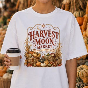 Könnte beinhalten: Ein weißes T-Shirt mit einem Grafikdruck und dem Text Harvest Moon Market. Das Design zeigt Illustrationen von Kürbissen, Zierkürbissen und Maiskolben in Holzkörben, ergänzt durch eine Mondsichel und einen Stern oberhalb des Schriftzuges.