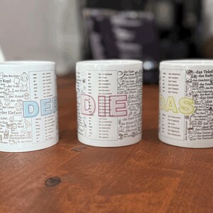 Op de afbeelding: Drie witte keramische mokken met Duitse woorden en illustraties. De mokken zijn gelabeld met "DER", "DIE" en "DAS" in grote, kleurrijke letters. Elke mok heeft een andere set Duitse woordenschat en tekeningen.