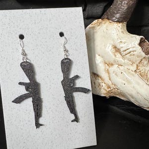 May include: A pair of silver dangle earrings with black silhouette gun charms. The earrings are displayed on a white card.