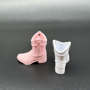 May include: Two miniature cowboy boots. One is light pink, and the other is white. The pink boot is fully formed, showing the details of the design. The white boot is a partial form, showing the inside of the boot.