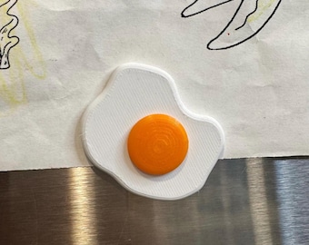 Può includere: Un magnete a forma di uovo fritto bianco stampato in 3D con un tuorlo arancione. Il magnete è su una superficie metallica, con un disegno colorato sullo sfondo. L'uovo è un oggetto decorativo divertente.