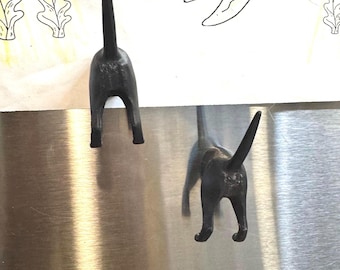 Könnte beinhalten: Zwei schwarze Katzen-Hintern-Magnete sind an einer Edelstahloberfläche befestigt. Die Magnete haben die Form eines Katzenhinterns, wobei der Schwanz nach oben zeigt. Die Magnete befinden sich in der Nähe einer bunten Zeichnung.