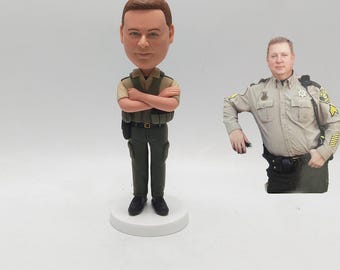 May include: A bobblehead figurine of a person in a sheriff's uniform, with arms crossed. The figurine is on a white base. A photo of a person in a sheriff's uniform is next to the figurine.