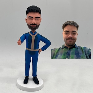 May include: A custom bobblehead figurine of a man in a blue jumpsuit with tan accents, giving a thumbs-up gesture. The figurine is on a white base, with a photo of the man in a plaid shirt next to it.