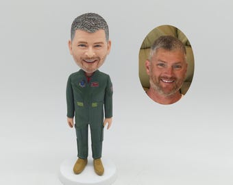 May include: A custom bobblehead figurine depicting a person with short gray hair and a beard, wearing a dark green flight suit with patches on the chest and sleeves. The figure stands on a round white base next to an oval photo of the person.