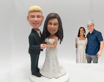 May include: A wedding cake topper featuring a smiling couple, the groom in a dark green suit and the bride in a white gown. The topper is on a white base. A photo of a smiling couple is in the background.