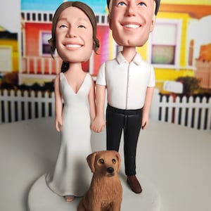 May include: A wedding cake topper featuring a bride in a white dress and a groom in a white shirt and black pants, holding hands. A brown dog sits in front of them. The figures are on a white base.