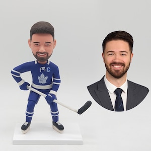 May include: A hockey player figurine in a blue and white uniform, with a hockey stick. The figurine has a smiling face and is on a white base. A portrait of a man in a suit is next to it.