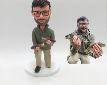 May include: A figurine of a man holding two fish, wearing glasses, a green hoodie, and khaki pants. The figurine is on a white base. A photo of a man holding two fish is next to the figurine.