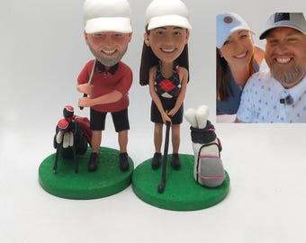 Op de afbeelding: Beeldjes van een man en een vrouw in golfkleding, elk met een golfclub en -tas. De man draagt een rood poloshirt, zwarte shorts en een witte pet. De vrouw heeft een zwart-rode ruitjestop, een zwarte rok en een witte pet.