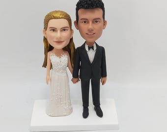 May include: A wedding cake topper featuring a bride and groom figurine. The bride wears a white wedding dress, and the groom is in a black tuxedo. They stand on a white base, holding hands. The bride has blonde hair.