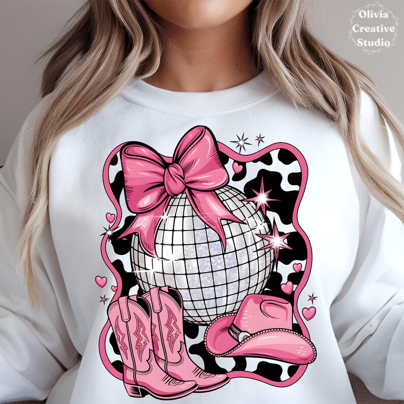 May include: A white sweatshirt featuring a pink disco ball, pink cowboy hat, and pink cowboy boots against a black and white cow print background. The disco ball is adorned with a pink bow.