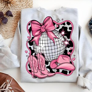 May include: A grey sweatshirt with a pink and white design featuring a disco ball, cowboy boots, and a cowboy hat. The design is set against a black and white cow print background.