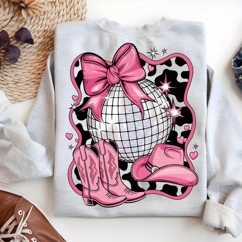 May include: A grey sweatshirt with a pink and white design featuring a disco ball, cowboy boots, and a cowboy hat. The design is set against a black and white cow print background.