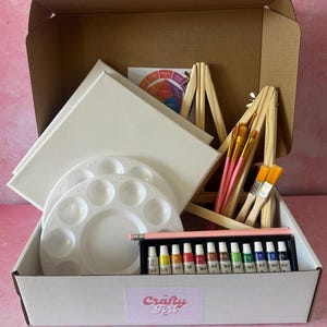 May include: A white box containing art supplies: paint tubes, paintbrushes, a colour wheel, two white canvases, and two white paint palettes. The box has the text "Crafty Girl" on the front.