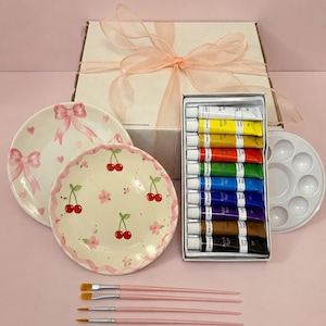 May include: A craft kit featuring two ceramic plates with hand-painted cherry and bow designs, a set of twelve acrylic paint tubes, a white plastic paint palette, and four paintbrushes with light pink handles, all packaged in a gift box.