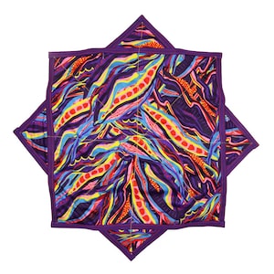 May include: Two square quilted pot holders with a vibrant abstract design. The pattern features flowing lines in shades of purple, blue, yellow, red, and orange. Each pot holder has a purple border and is quilted with a diamond pattern.