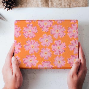May include: A rectangular box wrapped in vibrant orange paper featuring a repeating pattern of five-petaled pink flowers with yellow centers. The floral design is evenly spaced across the surface of the box, creating a bright and colorful appearance.