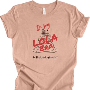 May include: A peach-colored t-shirt with a black and red graphic of a birthday cake with the text "In my Lola Era" and "Is that not allowed?!"