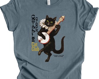 May include: Blue-grey t-shirt featuring a black cat playing a banjo. The cat wears a ruffled collar and has yellow eyes. Text on the shirt includes "Cripple Cat Club" and "Cool Cats Welcome."