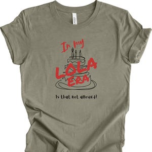 May include: A light green t-shirt with a black and red graphic of a birthday cake with candles and the text "In my Lola Era Is that not allowed?"