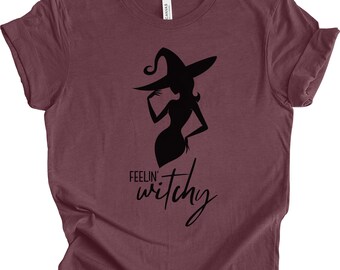 Può includere: T-shirt bordeaux con una silhouette nera di una strega con un cappello. Il testo "Feelin' Witchy" è scritto in corsivo sotto la grafica della strega. La maglietta è realizzata in materiale morbido.