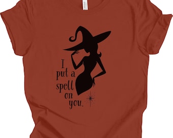 May include: Rust-colored t-shirt featuring a black silhouette of a witch wearing a hat. The text "I put a spell on you" is printed in black. A starburst graphic is below the text. The shirt is made of a soft material.