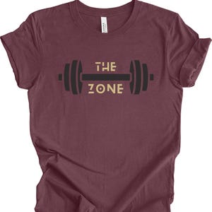 Könnte beinhalten: Burgunderfarbenes T-Shirt mit einem Hantel-Motiv. Der Schriftzug "THE ZONE" ist goldfarben. Das Shirt besteht aus einem weichen Material und hat hochgekrempelte Ärmel.