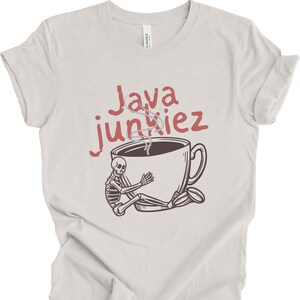 May include: A light beige t-shirt featuring a graphic of a skeleton hugging a large coffee cup with steam rising from it. Above the cup, the text Java junkiez is printed in a dark red, distressed font. The design is centered on the chest.