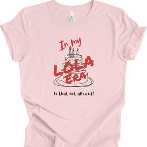 May include: Pink t-shirt with a black and red graphic of a birthday cake with candles and the text "In my Lola Era" and "Is that not allowed?"