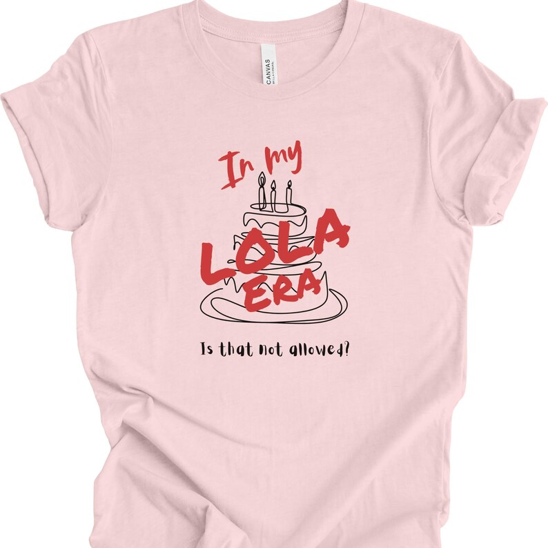 May include: Pink t-shirt with a black and red graphic of a birthday cake with candles and the text "In my Lola Era" and "Is that not allowed?"