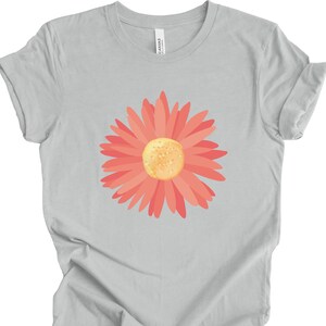 Puede incluir: Una camiseta gris claro con un gr&aacute;fico de margarita rosa. La flor tiene un centro amarillo.