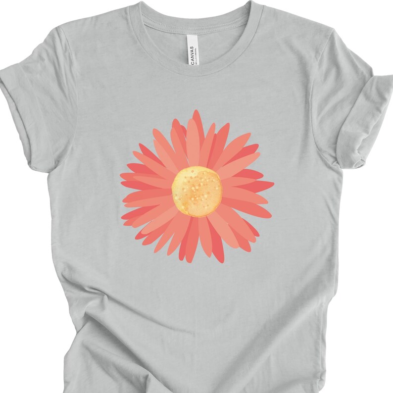Puede incluir: Una camiseta gris claro con un gr&aacute;fico de margarita rosa. La flor tiene un centro amarillo.