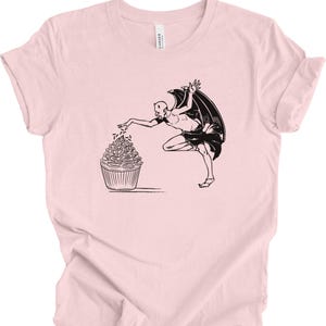 Può includere: Una maglietta rosa con un'illustrazione in bianco e nero di un diavolo che spolvera delle codette su un cupcake.