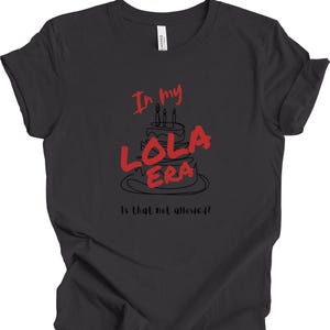 May include: A dark gray t-shirt with a black and white illustration of a birthday cake with the text "In my Lola Era" in red. The text "Is that not allowed?" is printed below the cake.